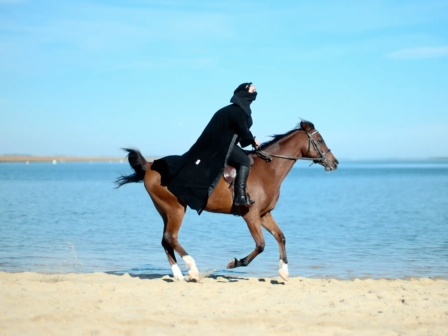 A rider in flowing black canters a bay horse through shallow water on the beach