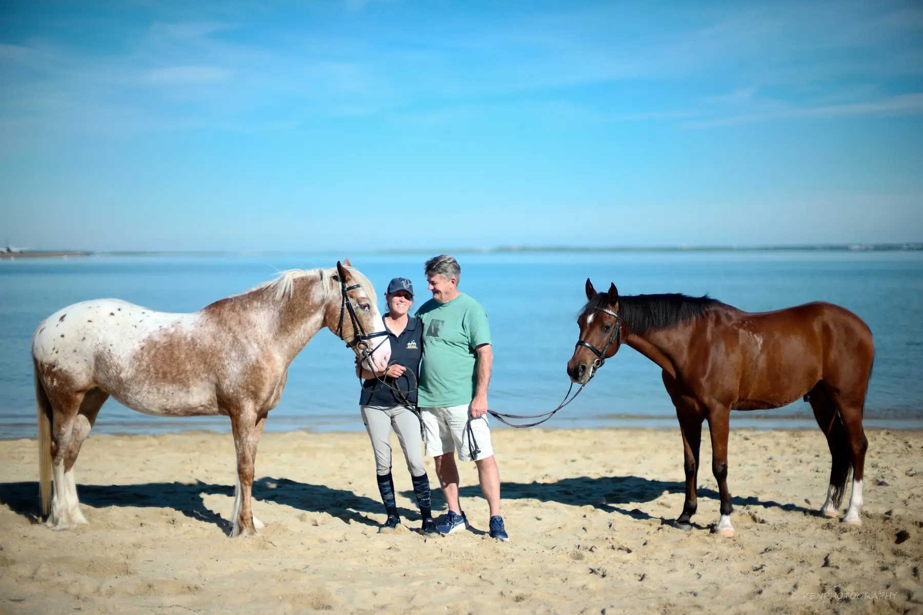 A family stands on the sand between an appaloosa and a bay horse, water behind them