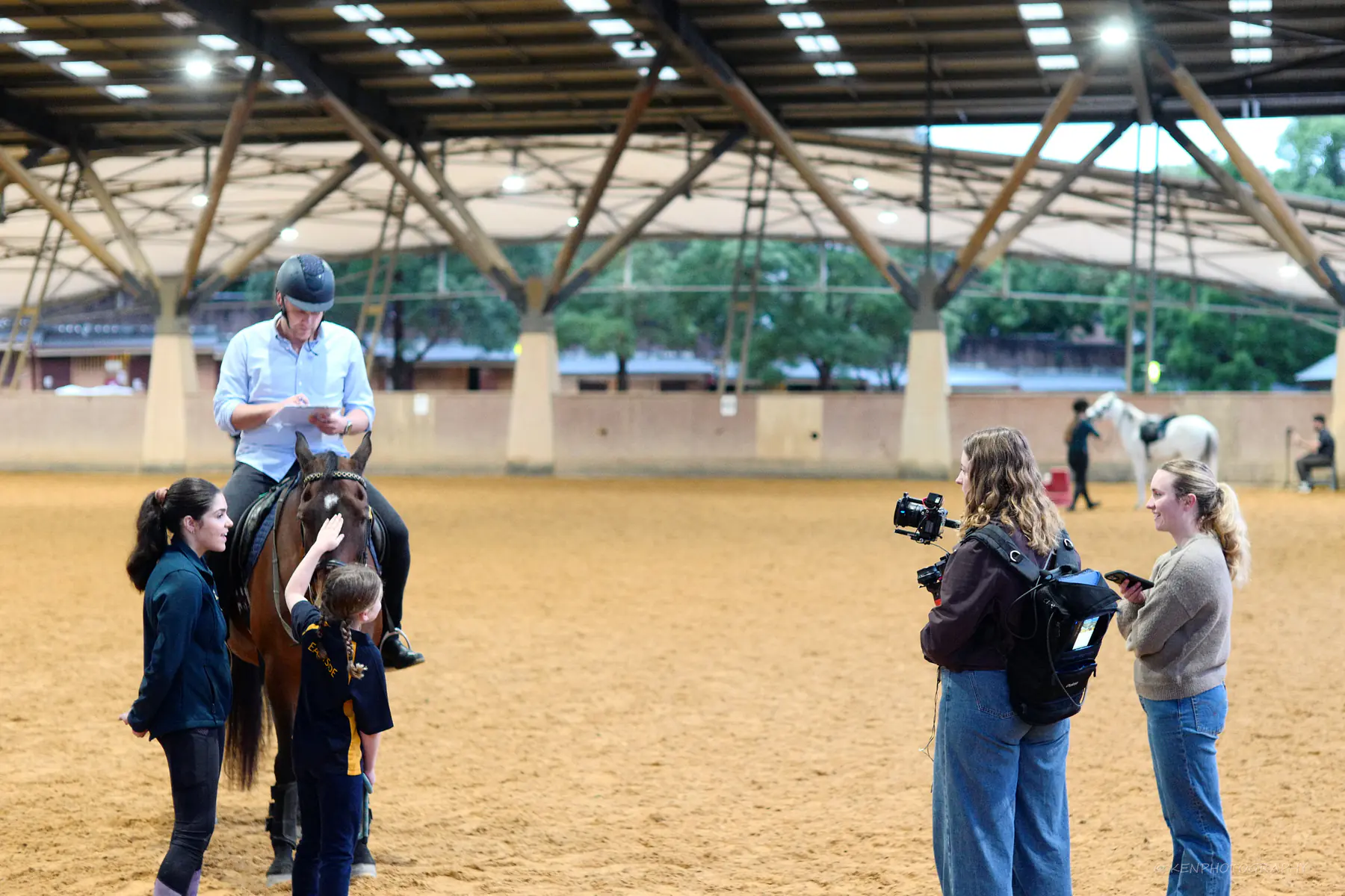 A film crew with camera works around a rider and a child leading a horse in the indoor arena