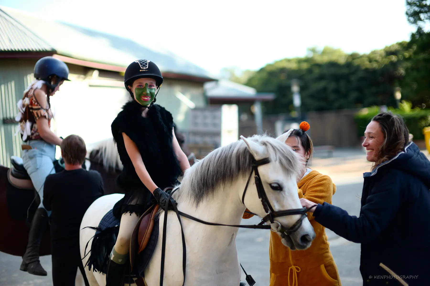 A child in face paint sits on a white pony held by a handler, Halloween costumes around them