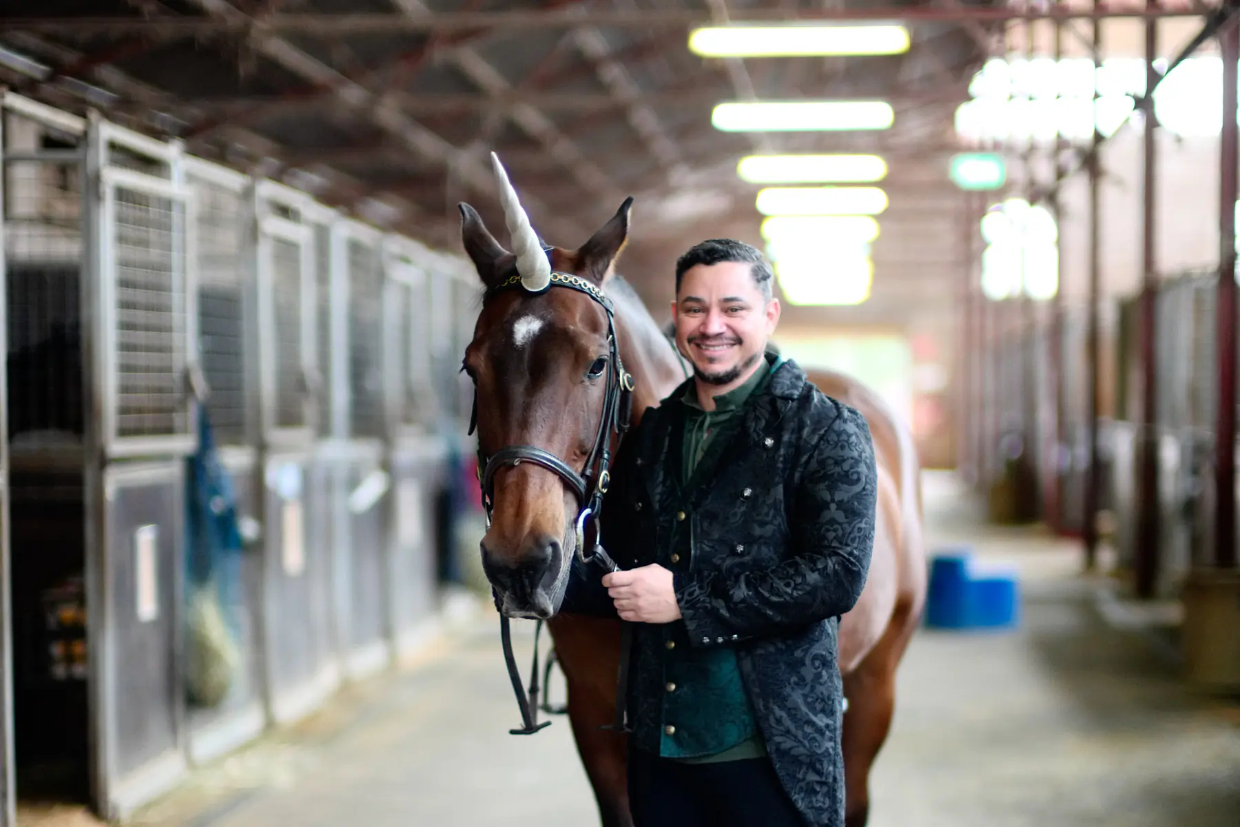 A man in a brocade jacket stands with a bay horse wearing a unicorn horn in the stable aisle